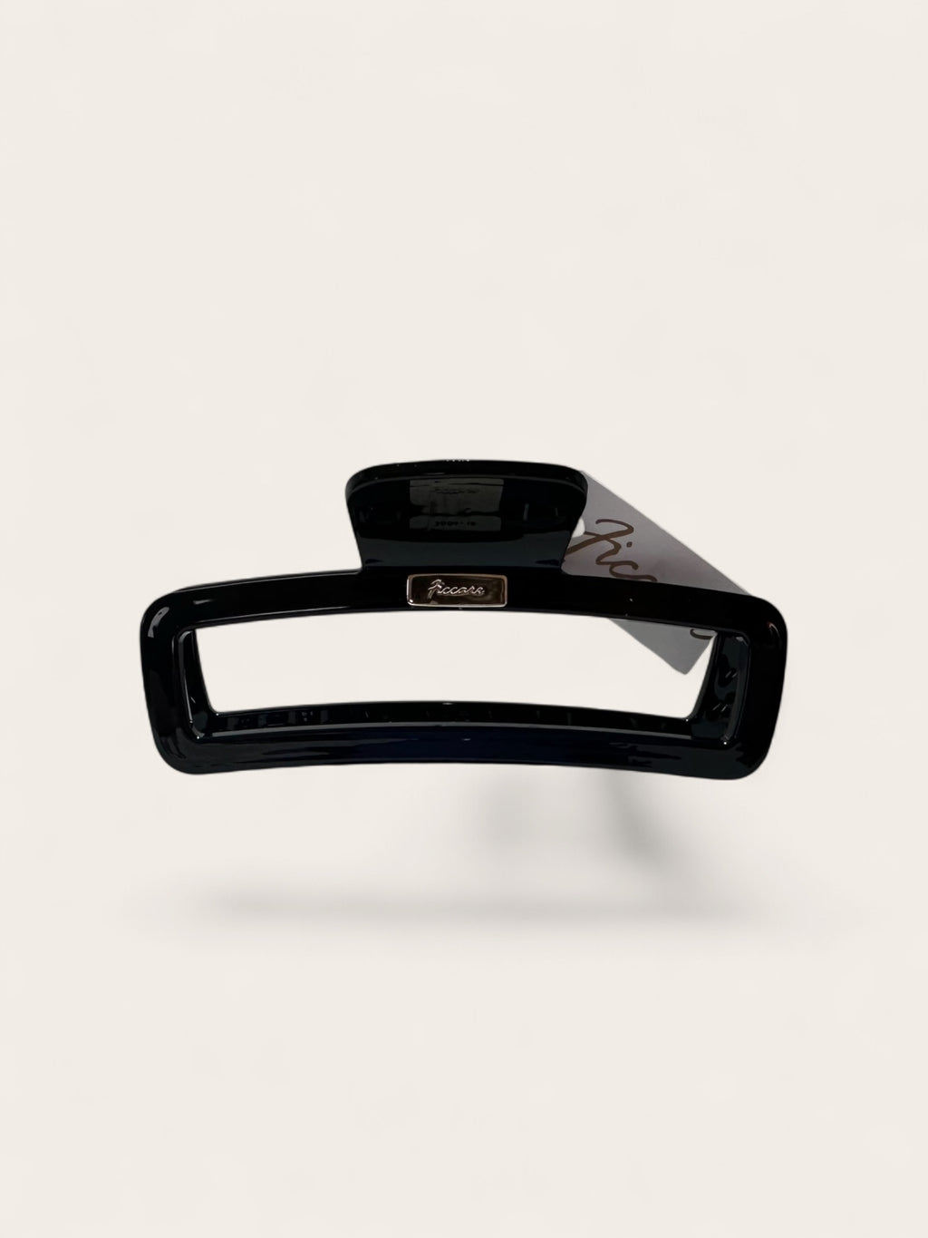 A glossy black hair claw clip with a small gold-tone brand plaque reading 'Ficcare' and a gray paper tag attached, shown on a clean off-white background.