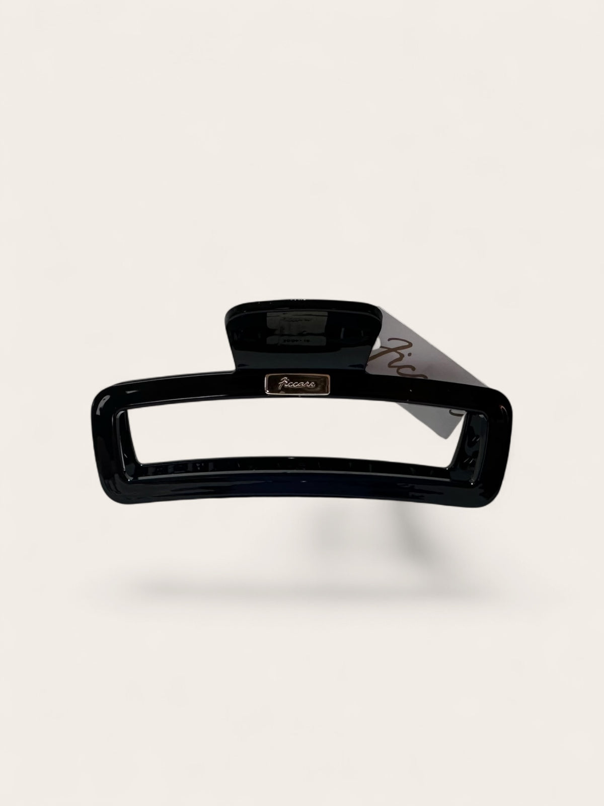 A glossy black hair claw clip with a small gold-tone brand plaque reading 'Ficcare' and a gray paper tag attached, shown on a clean off-white background.