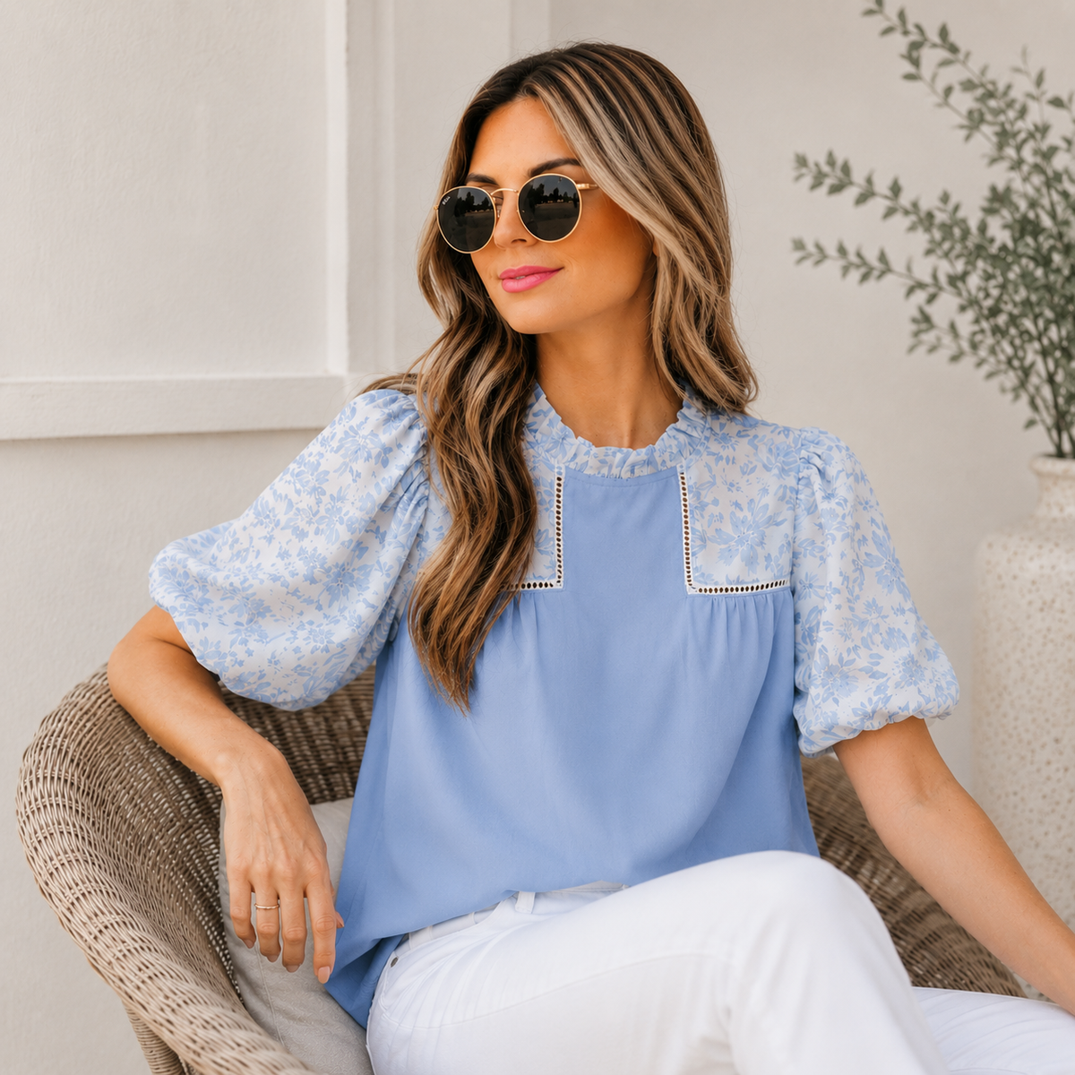 A woman wearing a light blue blouse with a delicate white floral print on the sleeves and collar, paired with white pants, sitting in a wicker chair.